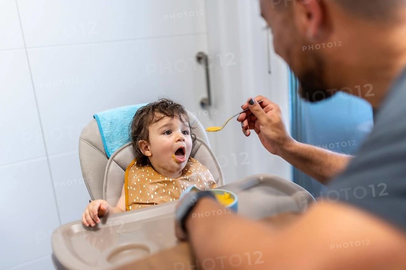 Father feeding vegetable puree to a happy baby in a high chair, celebrating family bonding and healthy eating habits during mealtime, representing early childhood nutrition and care