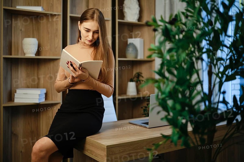 A woman sits on a desk in a modern office, reading notes. The room has wooden shelves with decorative items. A laptop and plants are visible in the background. Light enters through a window