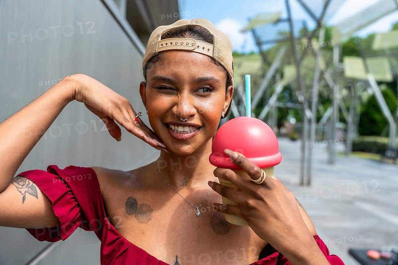 Young woman looking at camera, making a playful wink with a wide smile, wearing a cap and holding a refreshing sweet drink outdoors on a bright summer day