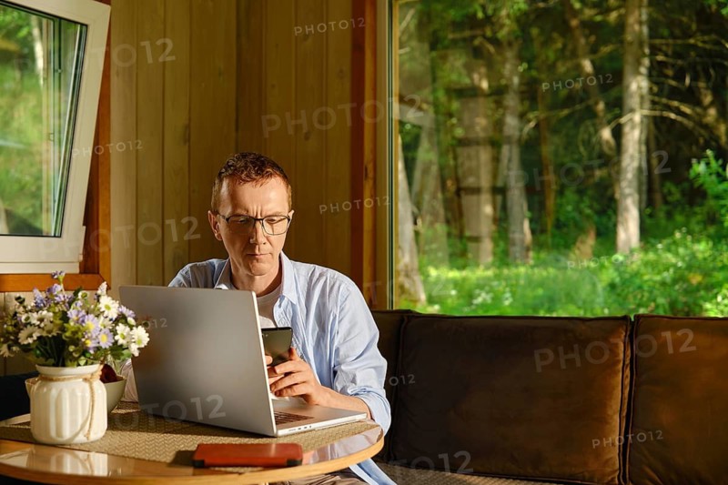 Middle-aged man works remotely from wooden country cabin. There is laptop on table in front of him and he is typing text message on his smartphone. Forest is visible through large windows