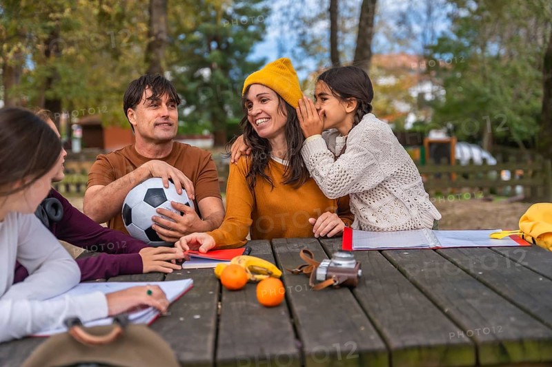 Family gathered at a wooden picnic table in a park as a daughter whispers to her mother, capturing warm bonding, trust, laughter and relaxed outdoor family time with snacks and a soccer ball