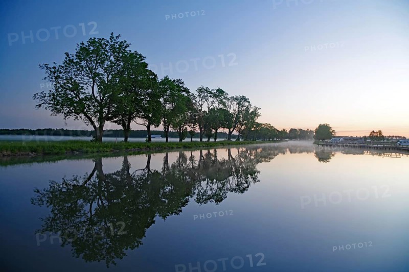 Trees are reflected in calm water under a morning sky, Hajstorp, Göta Canal, Västra Götalands län, Sweden