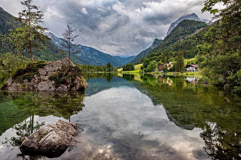 Clear mountain lake with rocks and tree in the foreground, framed by wooded alpine slopes, The Hintersee near Berchtesgaden in Bavaria