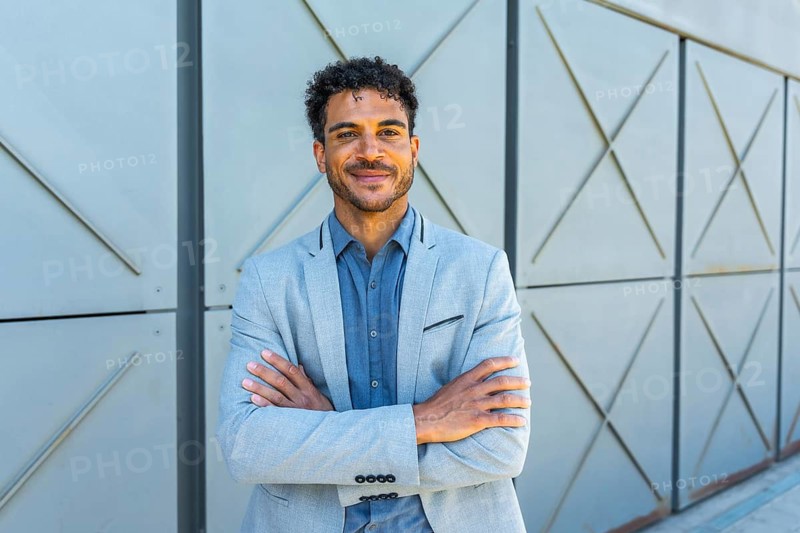 Young handsome man wearing a gray suit jacket and blue shirt, crossing arms and smiling at the camera, portraying confidence and professionalism in an urban setting