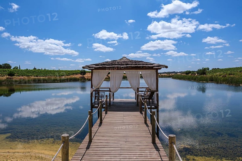 A wooden walkway with a pavilion stretches across a quiet, wide lake under a blue sky, Pedrógão, Beja, Portugal