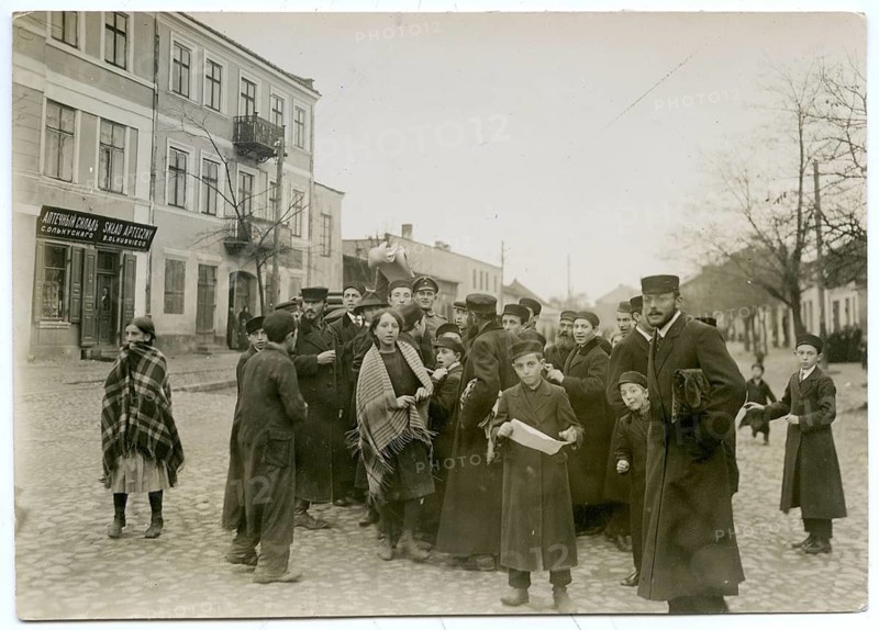 Students, pupils, handing out extra sheets, Jewish language, Jews, small town, Poland, German occupation, World War I, shops, a group of people in winter clothes standing together on a village street, historic atmosphere, photo Otto Haeckel, around 1916