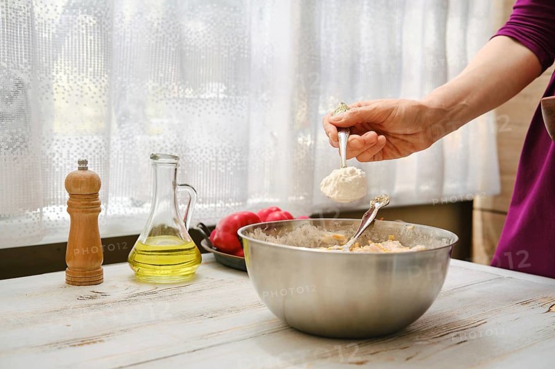 A person is measuring flour with a spoon over a mixing bowl. Fresh vegetables and cooking oil are on the counter. It is daytime and light comes through the window behind them