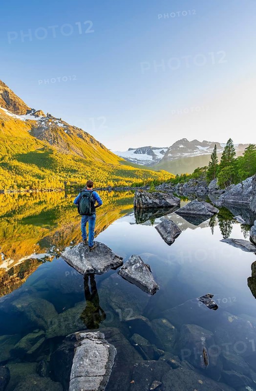 Young man standing on a rock in water, enjoying the landscape, picturesque mountain scenery in the Innerdalen high valley, mountain peaks reflected in Lake Innerdalsvatna, in the evening light at sunset, Trollheimen Mountain Area, Sunndal, Møre og Romsdal, Norway