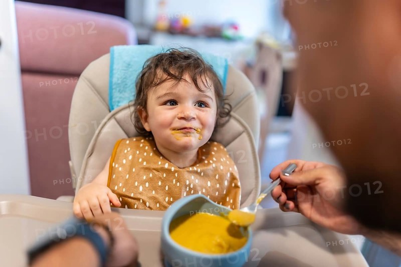 Cheerful baby with a messy face eating homemade vegetable puree in a high chair, smiling as a parent feeds themwarm moment of family bonding and healthy infant nutrition