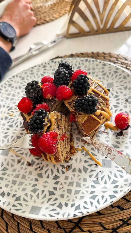 Stack of fluffy homemade pancakes topping with fresh raspberries, blackberries, and creamy peanut butter drizzle, enjoyed with a fork and knife on a patterned plate over a woven placemat