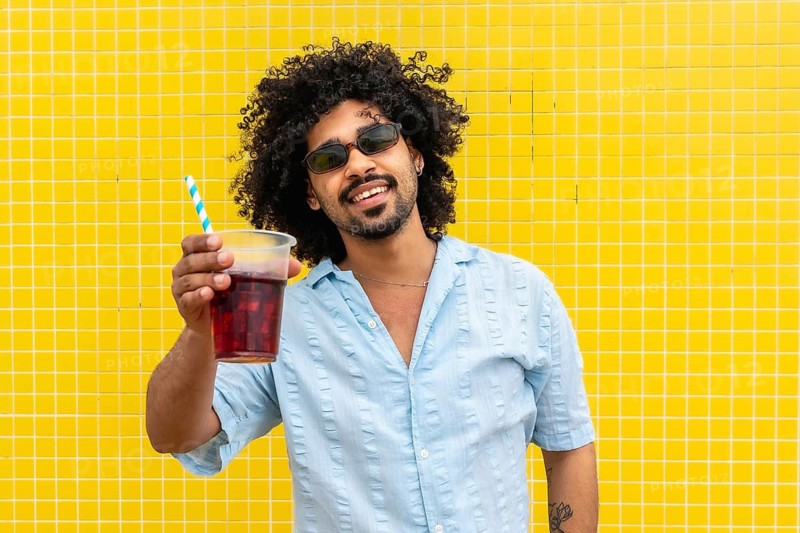 Young man with curly hair and sunglasses smiling while holding a cold drink with a straw, standing in front of a bright yellow tiled wall representing summer joy and freedom