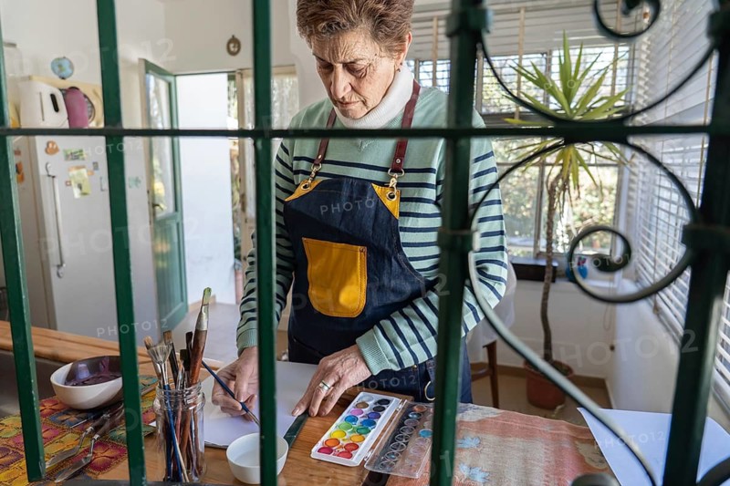Viewed through green decorative iron bars, an older woman in a yellow-pocket apron selects colors from a watercolor palette at her home studio table