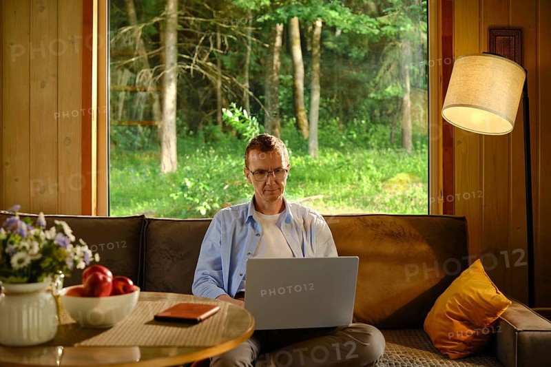Middle-aged man sits on couch in wooden cabin. He focuses on laptop while making online shopping and ordering delivery of goods. Red apples and notebook rest on table nearby