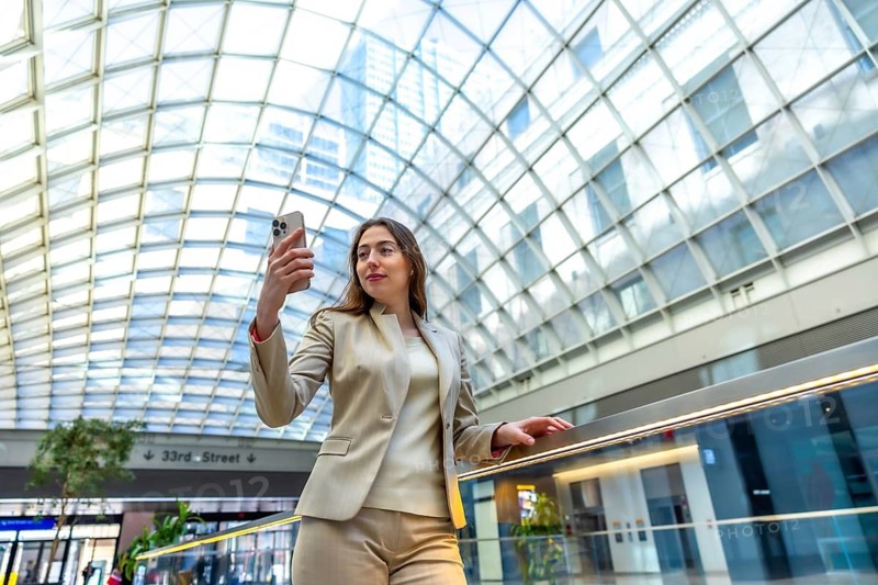Professional woman holding a smartphone, capturing a moment in a modern building with a distinct glass roof, reflecting an urban business lifestyle and connectivity