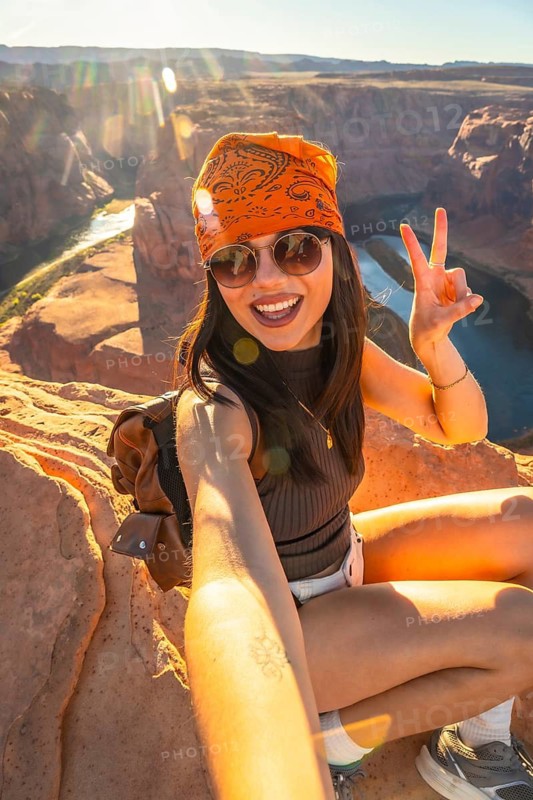 Young woman smiling and making a peace sign while taking a selfie on a rocky cliff overlooking the vast horseshoe bend canyon and colorado river during a sunny trip