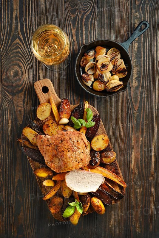 Roasted pork loin on a wooden board along with assorted vegetables. A glass of white wine sits beside the dish. A small skillet holds roasted onions