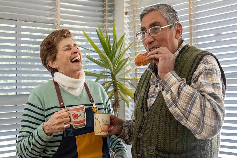 A senior woman bursts out laughing while holding two colorful ceramic mugs as her husband eats a croissant beside her, both sharing a joyful break in their bright home studio near an easel