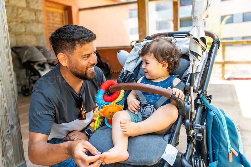 Cheerful father interacting with his smiling toddler son, who is sitting comfortably in a stroller with a colorful toy, capturing a beautiful moment of family bonding and parenting