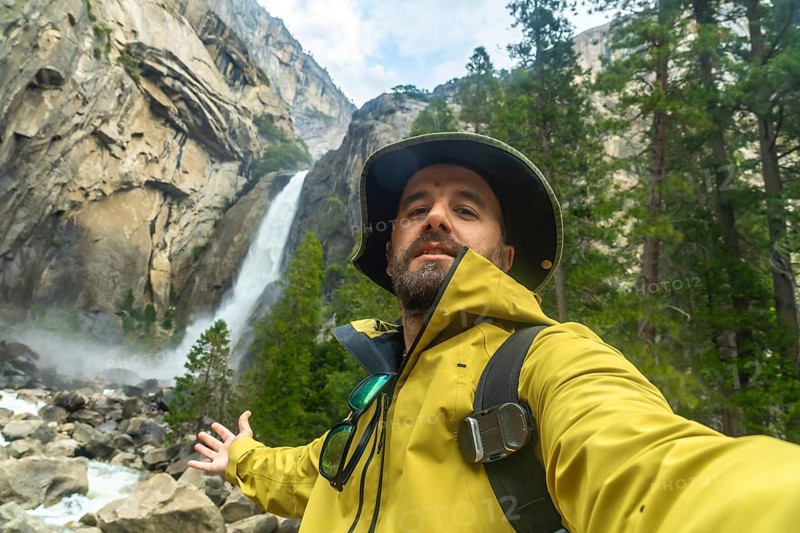 Man in a yellow jacket and hat taking a selfie, capturing himself enjoying the powerful lower yosemite falls waterfall and granite cliffs in yosemite national park, california