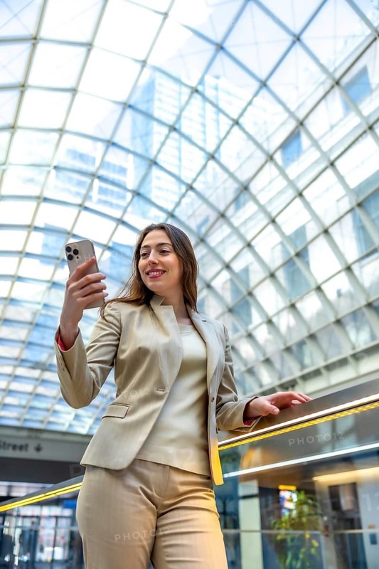 Businesswoman standing on an escalator in a modern glass domed building, smiling while holding a smartphone and engaging with technology in a bustling city environment