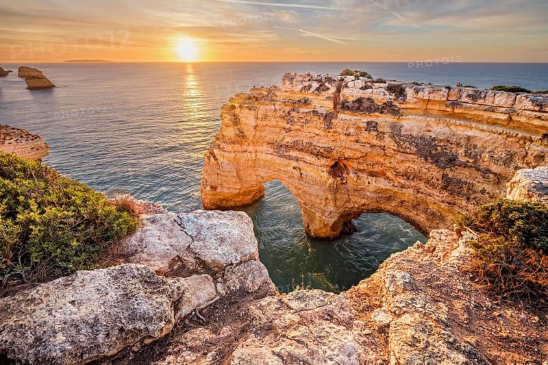 Sunrise view of Praia da Marinha beach in Algarve, Portugal, with a limestone sea arch, Atlantic water, warm sky, and rugged coastal cliffs