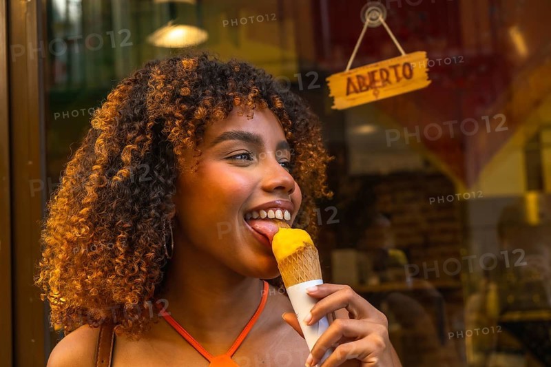 Young woman with beautiful curly hair smiling while licking a delicious scoop of yellow ice cream from a waffle cone on a warm summer day, standing in front of a shop with an aberto sign