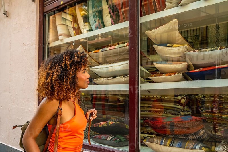 Young woman with curly hair window shopping, observing the display of colorful textile cushions and pillows inside a store showcasing home decor and accessories for sale