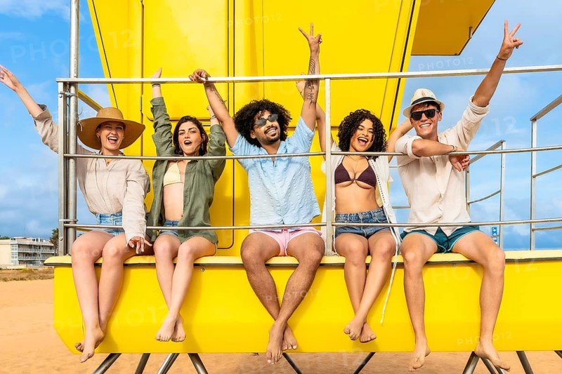 Group of diverse young people enjoying a joyful summer vacation on the beach, sitting cheerfully on a bright yellow structure with arms raised, embodying happiness, freedom, and friendship together