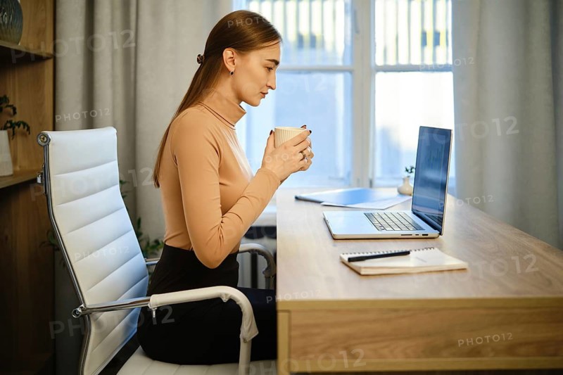 Woman sits at desk in her home office. She holds a cup and looks intently at her laptop screen