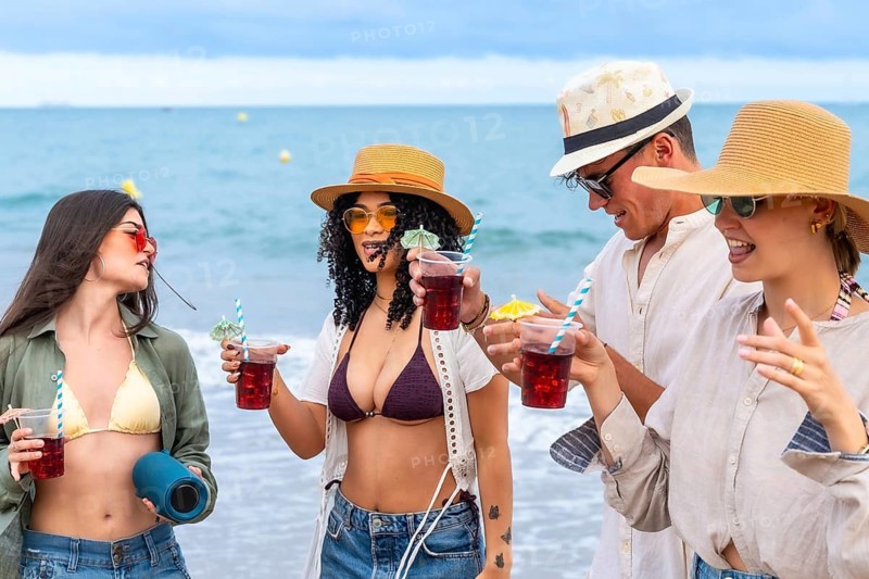 Young friends are celebrating summer vacation, enjoying refreshing drinks with straws and small umbrellas, having fun together on a sandy beach by the ocean