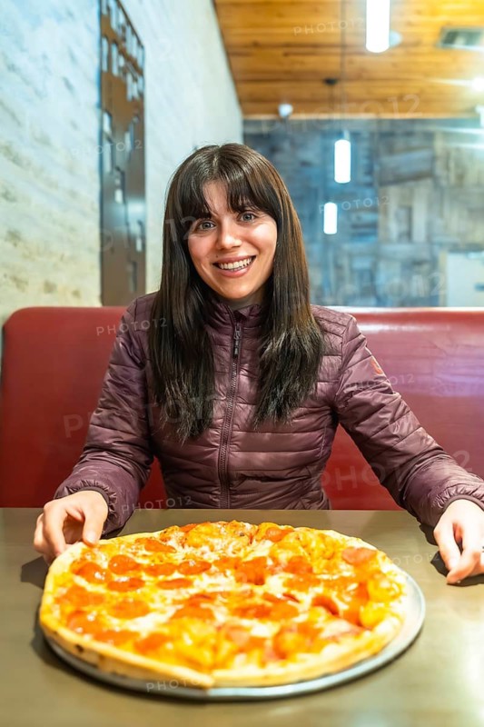 Young woman smiling while enjoying a hot pepperoni pizza in a casual restaurant booth after a day of travel, savoring cheesy comfort food and relaxed dining satisfaction