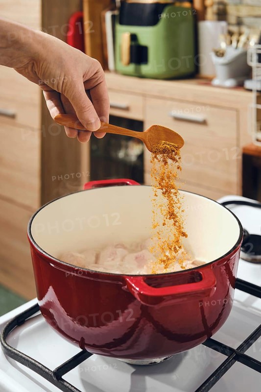 Hand adds spice from wooden spoon to cast iron pot on stove. Kitchen has cabinets and counter