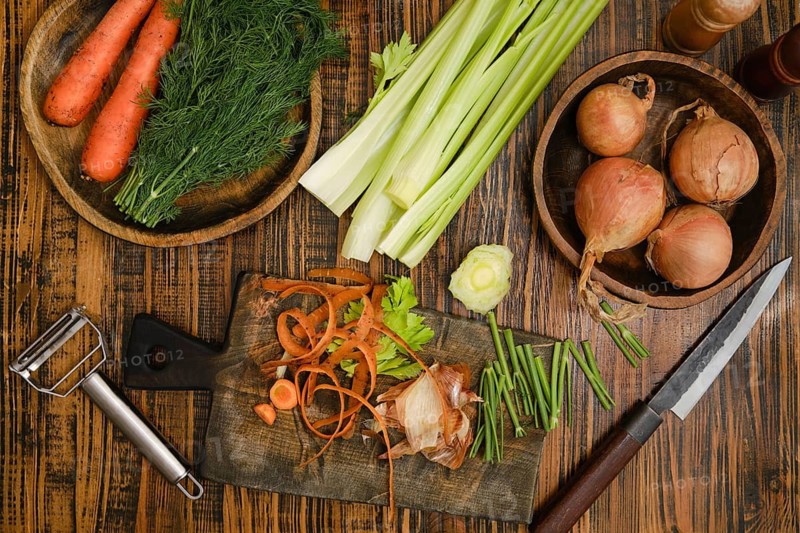 Carrots, celery, and dill are arranged on a wooden table. Vegetable peels are scattered on a cutting board
