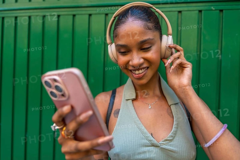 Young black woman with headphones smiling while looking at her smartphone, enjoying music and connecting with media on a green background
