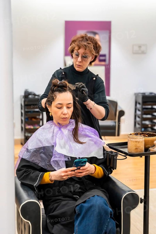 Professional hairdresser carefully applying hair dye to a young woman's hair, who is comfortably seated in a salon chair and using her smartphone during the treatment