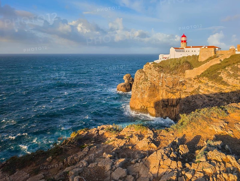 View of the Lighthouse of Cabo de Sao Vicente in Sagres, Algarve, Portugal, at sunrise with Atlantic ocean, cliffs, and golden foreground rocks