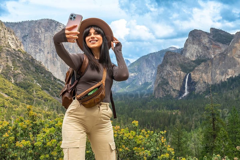 Woman standing at tunnel view in yosemite national park, holding a cell phone and taking a selfie with el capitan, bridalveil fall, and the valley in the background, enjoying a travel adventure