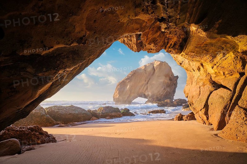 Sunset illuminates Penedo do Guincho, a rock arch at Praia da Santa Cruz, with ocean waves and sandy beach visible from the grotto