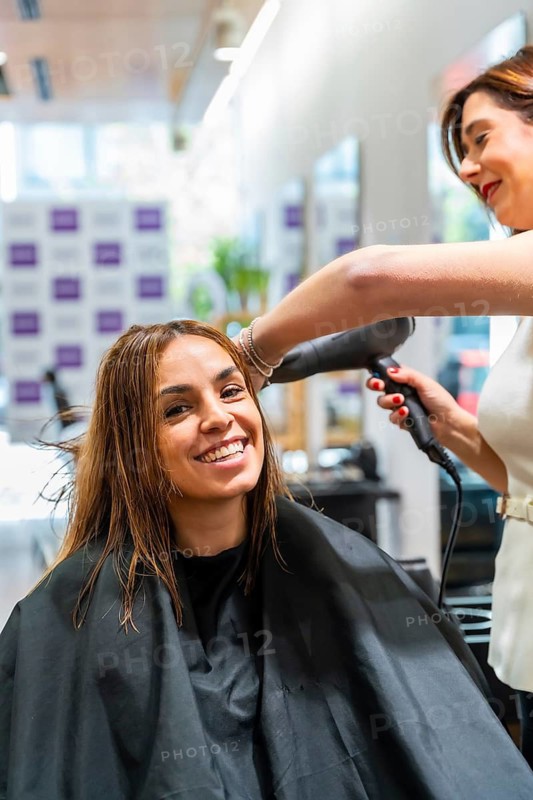 Smiling woman in salon chair receiving a professional blow dry from a hairstylist, enjoying pampering, hair care and styling for a fresh, healthy, cheerful look