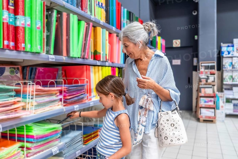 Grandmother and granddaughter shopping for stationery in a supermarket aisle, selecting colorful notebooks and binders for the back to school season and education