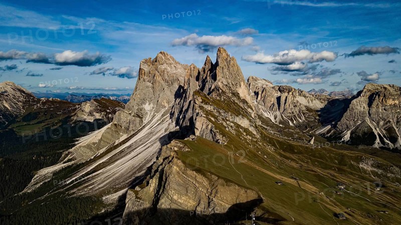 Impressive mountain formations under a clear sky with clouds, surrounded by green countryside, hiking trails wind their way through lush green valleys under the majestic peaks of the Seceda Mountains in the Italian Dolomites on a sunny summer day