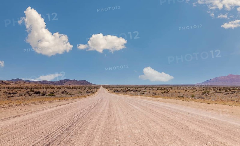 Endless dusty road through dry desert under blue sky with scattered clouds, Namibia