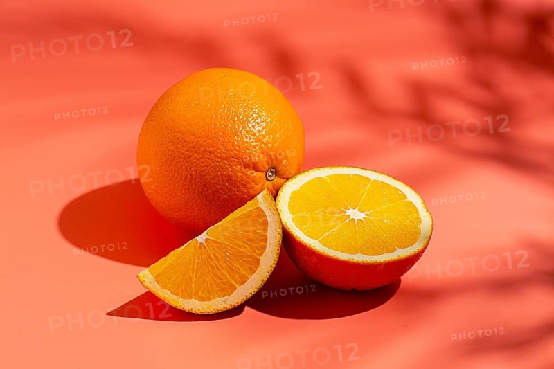 On the coral surface, next to two neatly sliced slices, there is a whole orange. Sunlight casts soft shadows, emphasizing the freshness and bright color of the fruit
