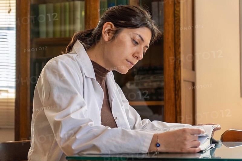 Young female scientist in white lab coat reading intently from a large open book at a glass desk, with an antique wooden bookcase full of books in the warm background