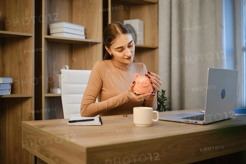 A woman sits at a wooden desk in a home office. She holds a piggy bank in her hands and gazes at it. A laptop and a mug are on the desk. Shelves with books are in the background