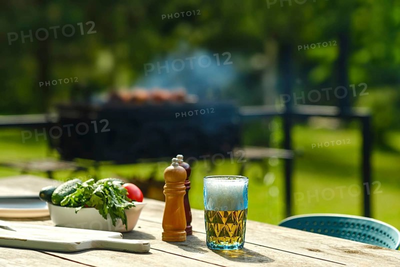 Glass of cold beer on wooden table next to fresh vegetables in backyard. Smoke rises from barbecue grill in background filled with cooking food