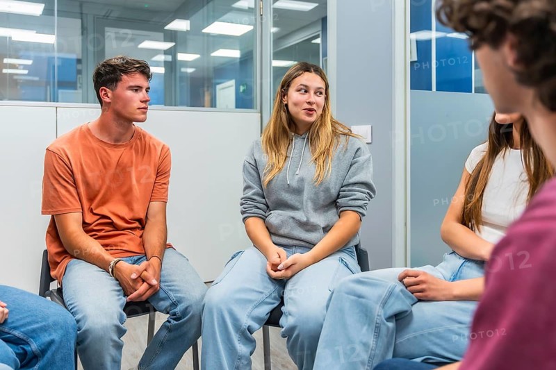 Young adults sitting in a circle, listening to a peer speaking during a group therapy session, fostering open communication, empathy, and providing support for mental health and personal growth