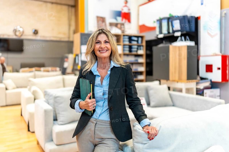 Confident saleswoman holding a tablet, standing in a furniture store showroom with new sofas, offering design advice and excellent customer service for home improvement