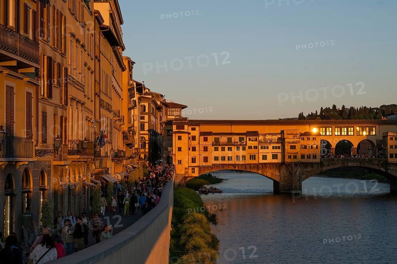 People stroll along the Arno River at sunset, warm evening light falls on the historic Ponte Vecchio segmental arch bridge and is reflected in window panes, evening mood, old town, Florence, Tuscany, Italy