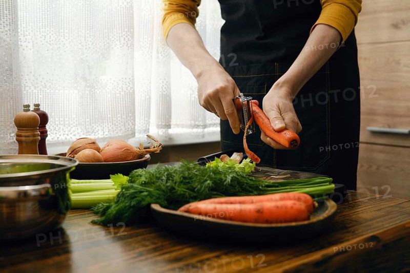 A person is peeling a carrot while standing at a wooden table filled with fresh vegetables. Celery, onions, and carrots are arranged on the table in a bright kitchen with natural light